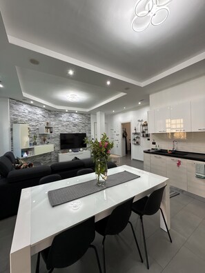 A modern living room with a white dining table, black chairs, a black sofa, a TV, and a stone-patterned wall.