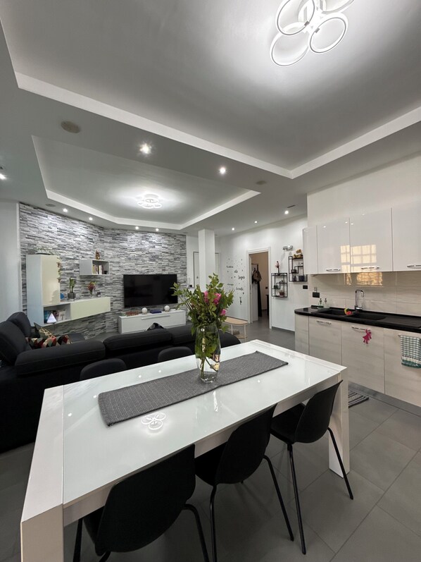 A modern living room with a white dining table, black chairs, a black sofa, a TV, and a stone-patterned wall. - Lucky's Nest - Apartment in the Heart of Asti (Asti)