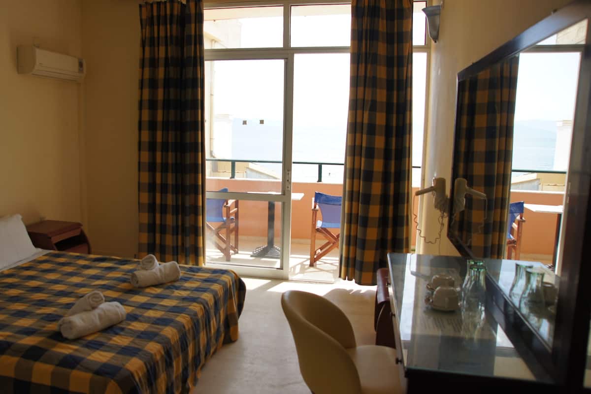 Habitación de hotel con cama, escritorio, silla, ventana con cortinas y balcón con sillas.