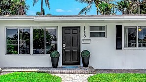 Un bâtiment blanc avec une porte noire, portant le numéro 1230, et deux grands plants en pot placés devant l’entrée.