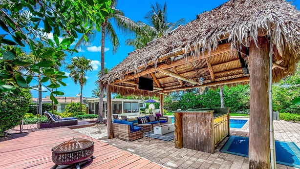 Une terrasse en bois surmontée d’une structure à toit de chaume, une piscine, des chaises longues et un espace bar.