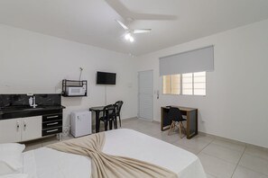Quarto de hotel com cama, escrivaninha, cadeira, televisão, ventoinha de teto e janela.