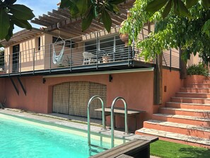 Ein Schwimmbecken mit Holzterrasse, einer überdachten Sitzgruppe im Freien und einer Treppe zu einer zweiten Ebene.