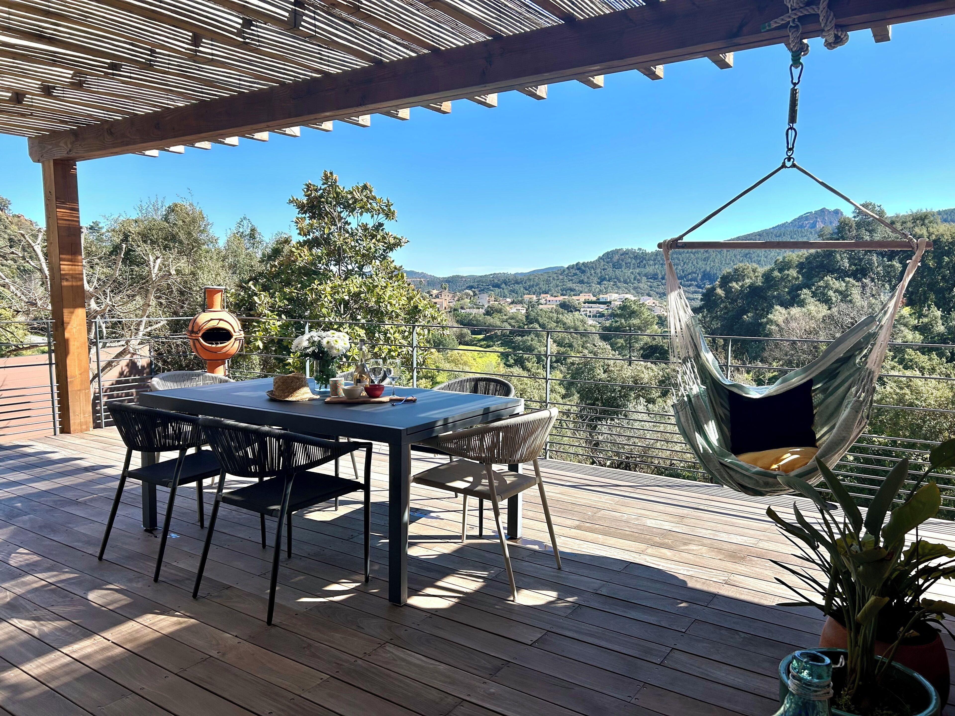 Um deck de madeira com mesa e cadeiras, uma cadeira suspensa e vista para um vale com árvores e edifícios.