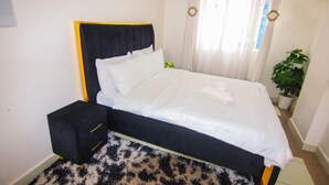 Una cama con cabecera negra y ropa de cama blanca, una mesita de noche negra con cajones, una planta en maceta y cortinas de gasa.