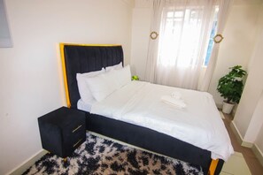 A bed with a black headboard and white bedding, a small black nightstand with drawers, a potted plant, and sheer curtains. - Bosa Homes (Nairobi)