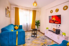 A living room with a blue sofa, a TV mounted on the wall, a small table, and a cabinet with a plant on top. - Bosa Homes (Nairobi)