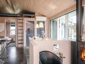 Un intérieur moderne avec un plafond en bois, un espace salon central doté d’une table et de chaises, une cheminée et de grandes fenêtres.