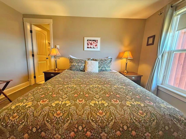 Une chambre à coucher avec une couvre-lit à motifs floraux, deux lampes de chevet, un tableau encadré au mur et une fenêtre avec des rideaux.