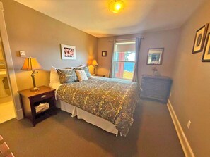 A bedroom with a bed, nightstands, a dresser, a window with curtains, and a framed picture on the wall. - Oceanfront Suite with Relaxing Tub & Private Bath - Rm 6 (Gabriola)