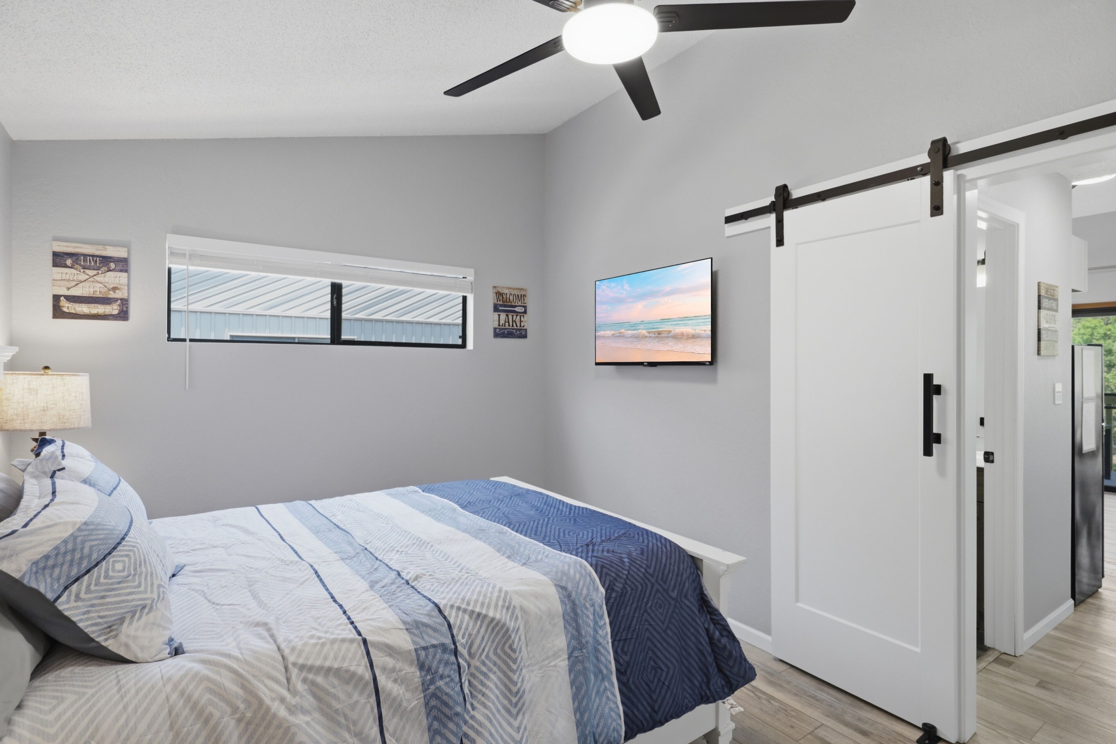 Een slaapkamer met een bed, een plafondventilator, een raam met jaloezieën, een aan de muur bevestigde televisie en een deur.