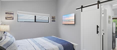 A bedroom with a bed, a ceiling fan, a window with blinds, a wall-mounted TV, and a door.