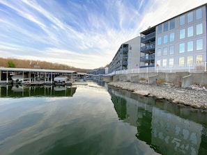 A waterfront property with a dock, a building with balconies, and a calm river. - H Lakefront Property, Remodeled, Boat Slip, Patio Views, Pool, Ideal Location! (Osage Beach)