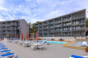 A poolside area with lounge chairs and umbrellas, a pool, and a multi-story building with balconies. - H Lakefront Property, Remodeled, Boat Slip, Patio Views, Pool, Ideal Location! (Osage Beach)