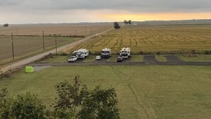 Paisaje rural con campos, un camino de tierra y varias autocaravanas estacionadas en un claro.