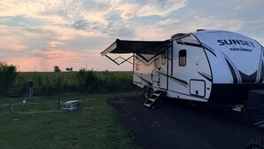 Una caravana blanca con el rótulo 'Sunset Trail Super Lite' estacionada en un camino rural, con un campo cubierto de césped y un cielo nublado al fondo.
