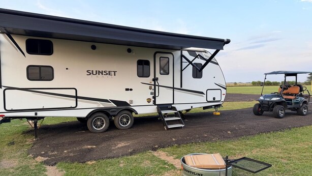 Una caravana blanca con la marca 'SUNSET TRAIL', un carrito de golf y un contenedor metálico etiquetado 'COUNTYLINE' en un camino de tierra.