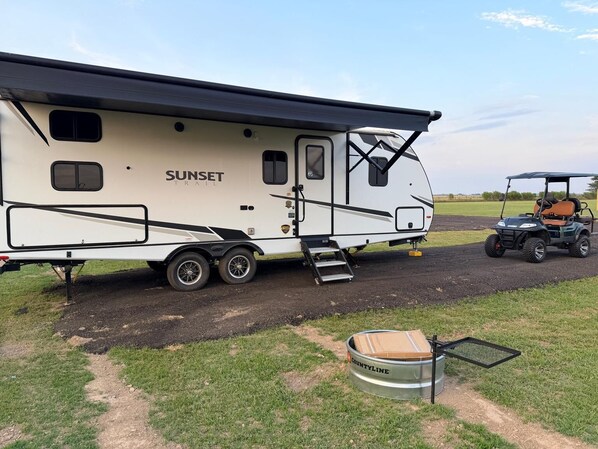 A white RV with 'SUNSET TRAIL' branding, a golf cart, and a metal container labeled 'COUNTYLINE' on a dirt path.