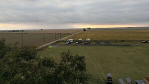 Paisaje rural con campos, vehículos estacionados y cielo nublado.