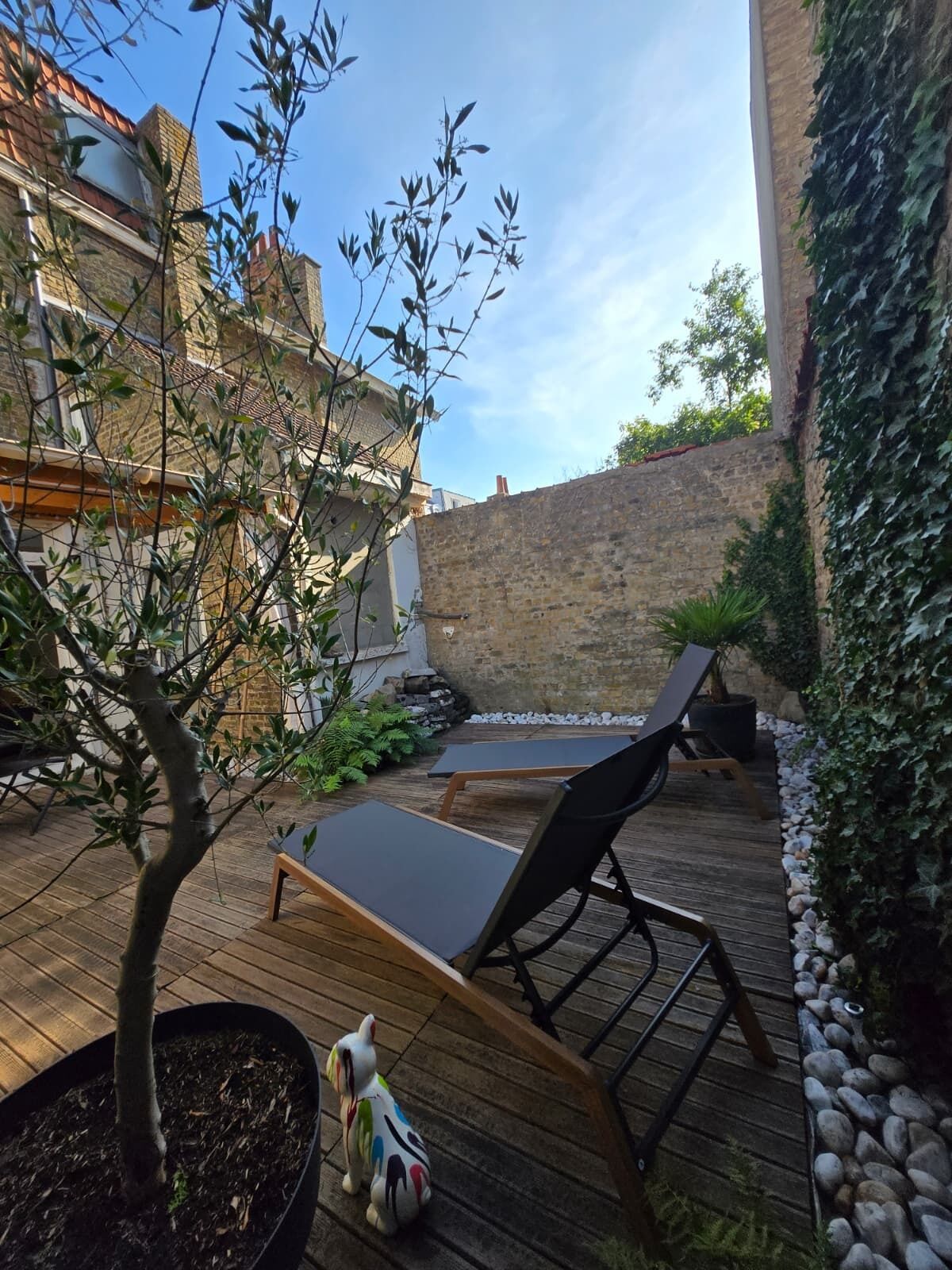 Una terrassa de fusta amb gandules, un arbre petit i una estàtua decorativa d'un gos.