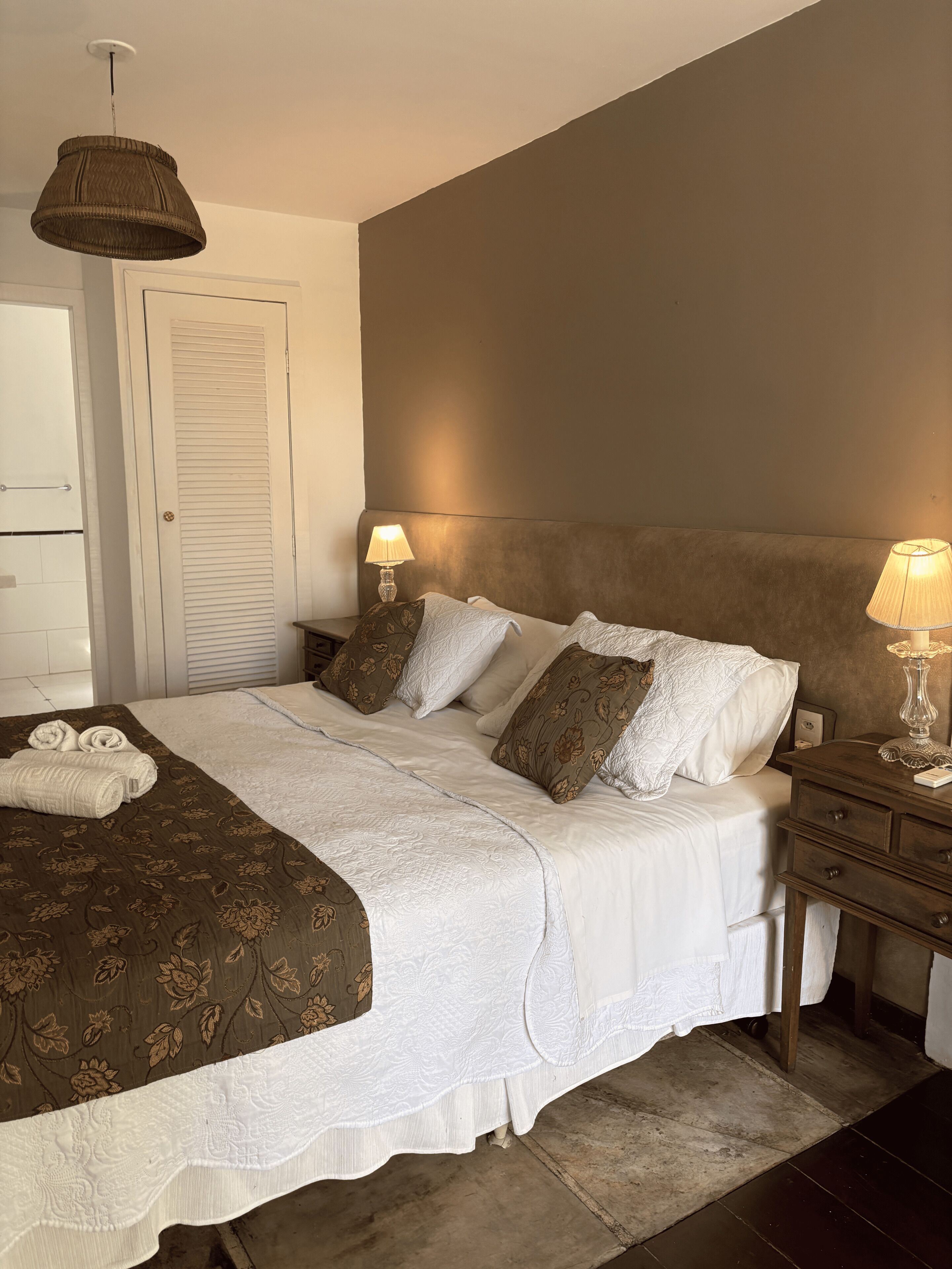A neatly made bed with white linens, two bedside lamps, a brown headboard, and a door with a towel rack.