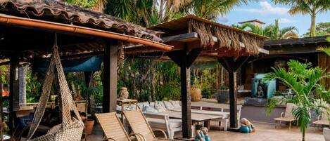 A covered outdoor seating area with wicker furniture and hanging chairs, surrounded by lush greenery and palm trees.