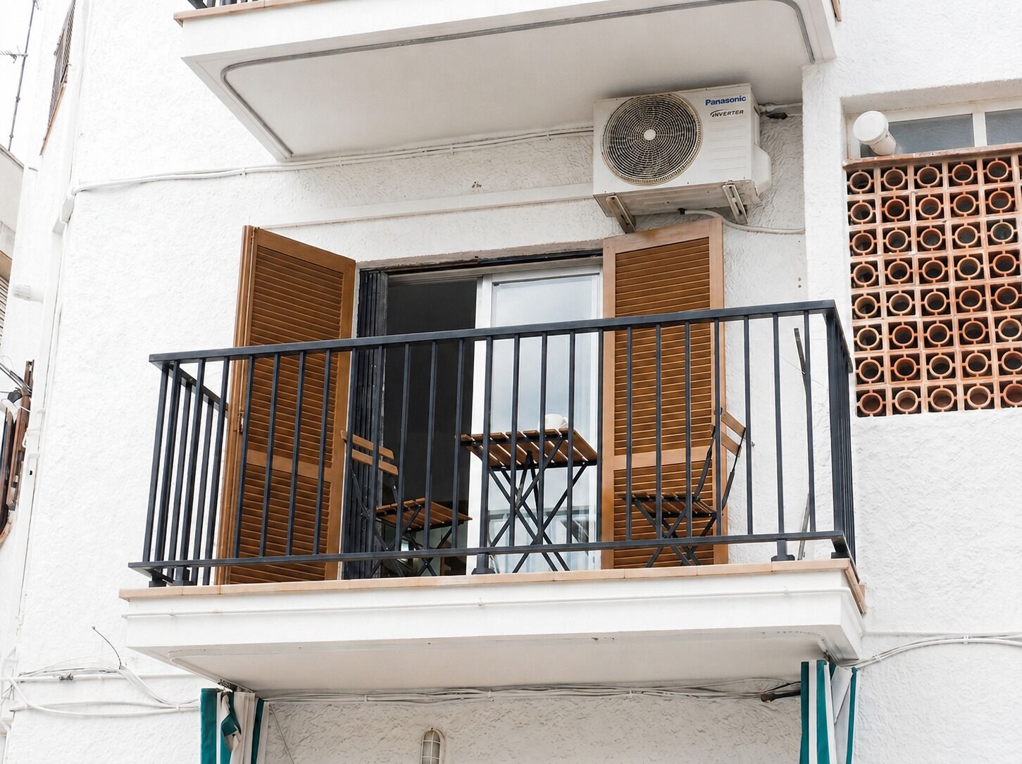 A balcony with a table and chairs, an air conditioning unit, and a window with wooden shutters.