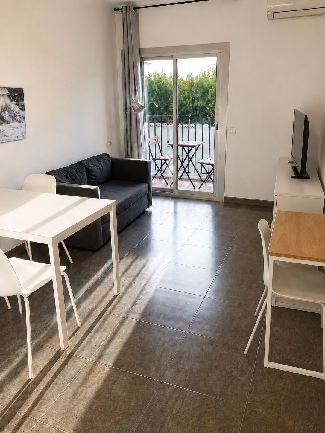 A modern living room with a sofa, a dining table, and chairs. There is a balcony with a view of greenery.