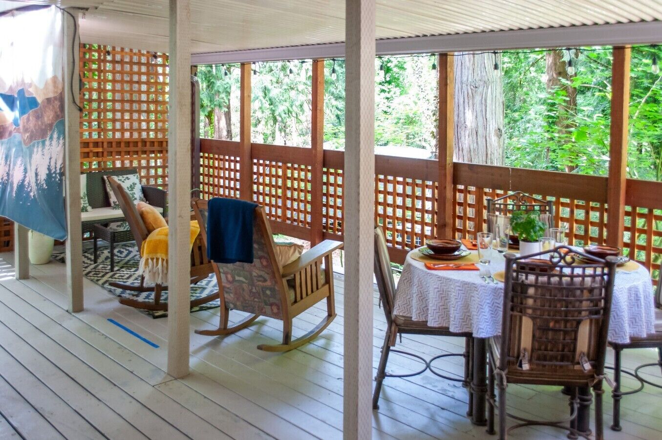An outdoor patio with wooden furniture, a table set for dining, and a view of greenery through a lattice structure.