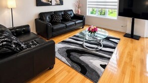 Een woonkamer met een zwarte leren bank, een glazen salontafel, een tapijt met zebraprint, een schilderij aan de muur, een raam met jaloezieën en een tv op een dressoir.