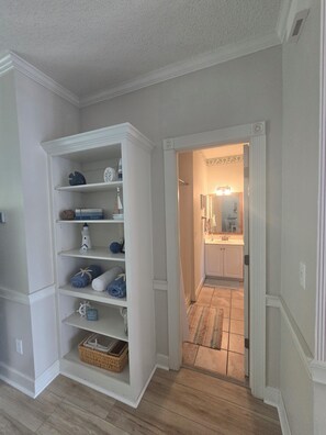 A hallway with a white built-in shelving unit, a wooden floor, and a view into a bathroom with a sink and mirror. - Arcadian Shores 2BR/2BA Condo Sleeps 6 • Pool • Balcony • Near Beach (Myrtle Beach)