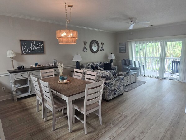 A dining area with a wooden table and chairs, a chandelier, and a sofa with a floral pattern. - Arcadian Shores 2BR/2BA Condo Sleeps 6 • Pool • Balcony • Near Beach (Myrtle Beach)