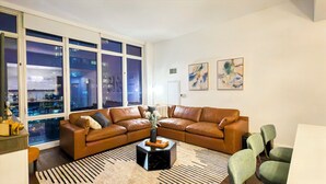 Un salon moderne avec un canapé d’angle en cuir, une table basse noire et une vue sur la ville grâce à de grandes fenêtres.