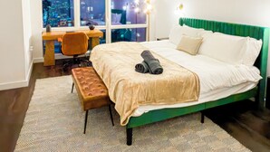 Une chambre avec vue sur la ville, un lit recouvert d’un tissu vert, un bureau et un tapis à motifs.