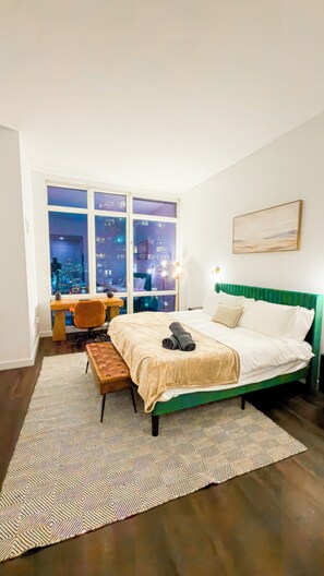 A bedroom with a city view, a green upholstered bed, a desk, and a patterned rug. - Luxury Midtown West High Rise with Skyline Views (New York)