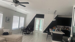 Una sala de estar moderna con sofá, mesa de comedor y sillas, una cocina con televisor, y una escalera que lleva a otro nivel.