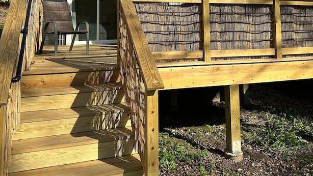 Eine Holzterrasse führt zu einer Hütte mit Fenster und Tür, umgeben von Bäumen.