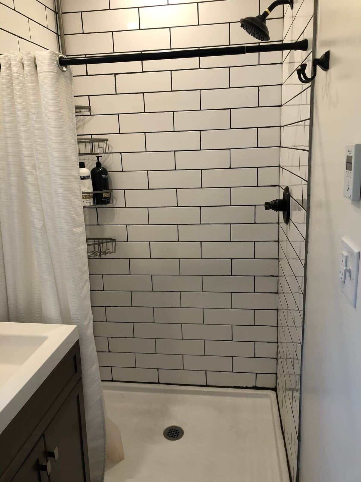 A bathroom with white subway tiles, a shower with a black rain showerhead, a white curtain, and a wall-mounted soap dispenser.