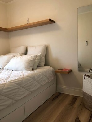A bedroom with a bed, a wooden shelf, a bedside table, a mirror, and a basket. - Charming & Quaint 1 bed in Historic Uptown! (Kingston)