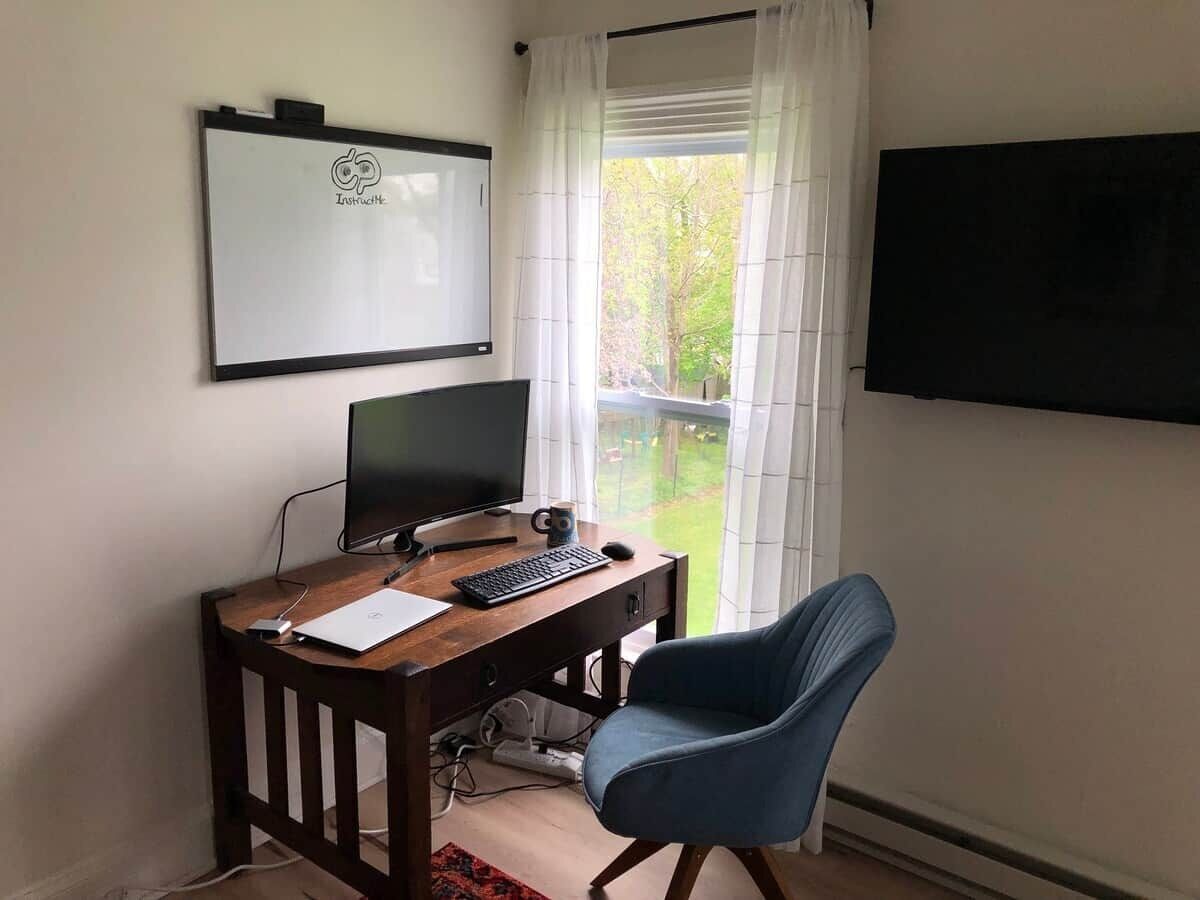 Una habitación con escritorio, computadora, silla, televisor y ventana con cortinas.