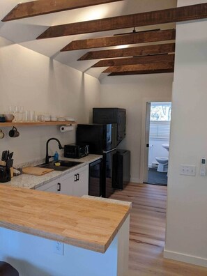Cocina compacta con encimera de madera, un fregadero y un microondas. Hay una puerta que da a un baño con inodoro y lavabo.