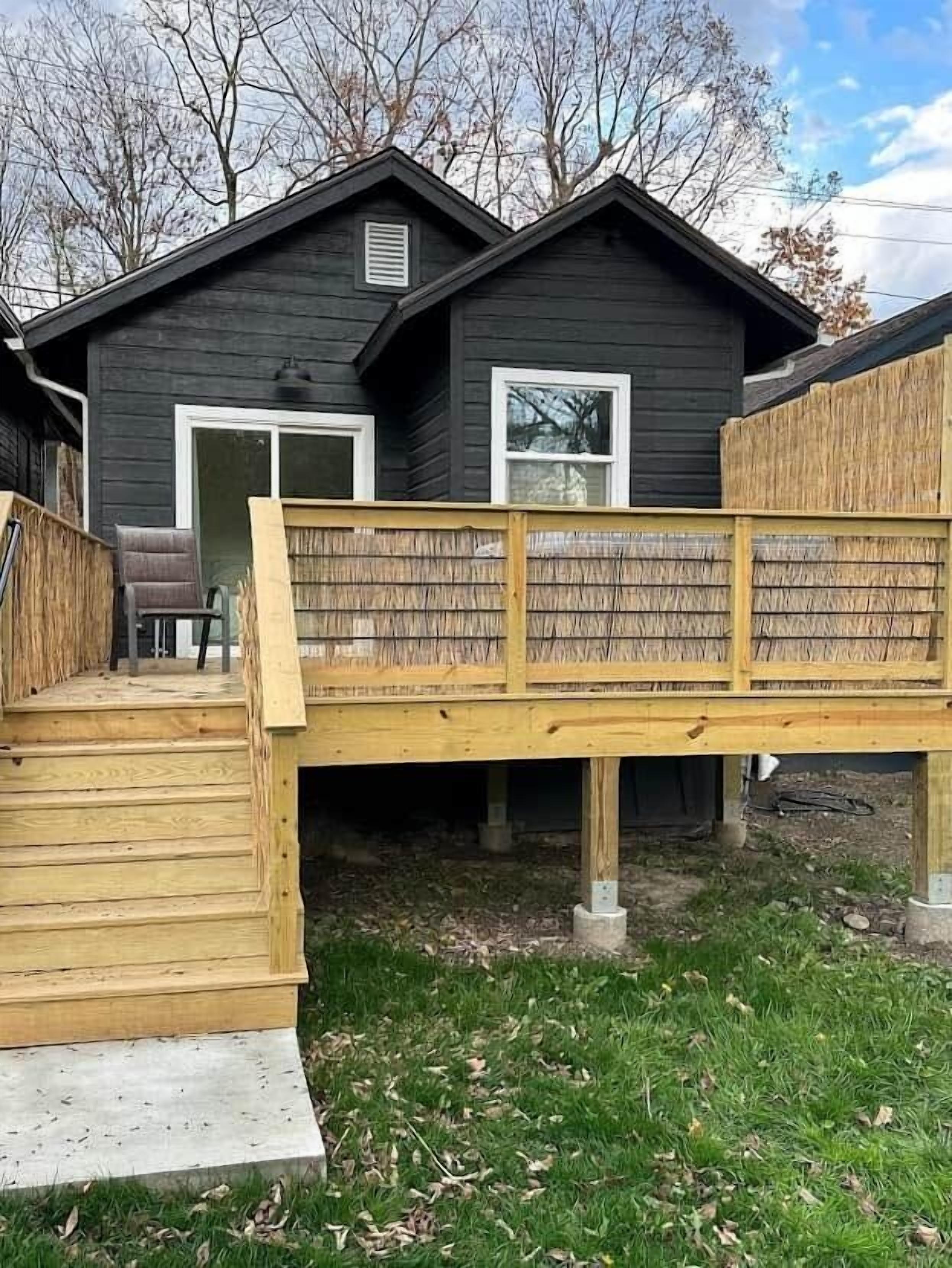 Een houten terras leidt naar een huis met een donkere gevel en witte ramen.