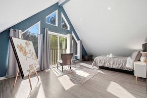 A bedroom with a sloped ceiling, a bed, a chair, a painting on easel, and a view of trees through large windows.