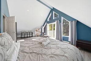 A bedroom with a large bed, a small table with plants, a painting, and a view through a window.