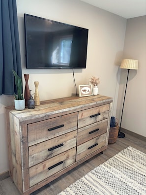 Un meuble en bois avec des tiroirs, un téléviseur fixé au mur, une lampe sur pied et un tapis à motifs.