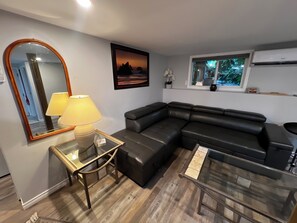 A modern living room with a sectional sofa, a glass coffee table, a lamp, a mirror, and a framed picture on the wall. - Trails End Beach House 1 bedroom plus cabin bedroom (Ladysmith)