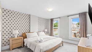Ein Schlafzimmer mit einem Bett, Nachttischen, einem Schreibtisch, einem Fenster mit Vorhängen und einer Wand mit Muster.