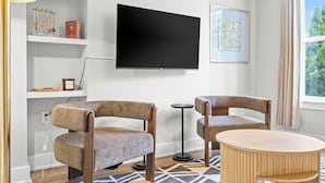 Ein modernes Wohnzimmer mit Fernseher, zwei Sesseln, einem runden Holztisch, einem gemusterten Teppich und einem Bücherregal mit Dekoration.
