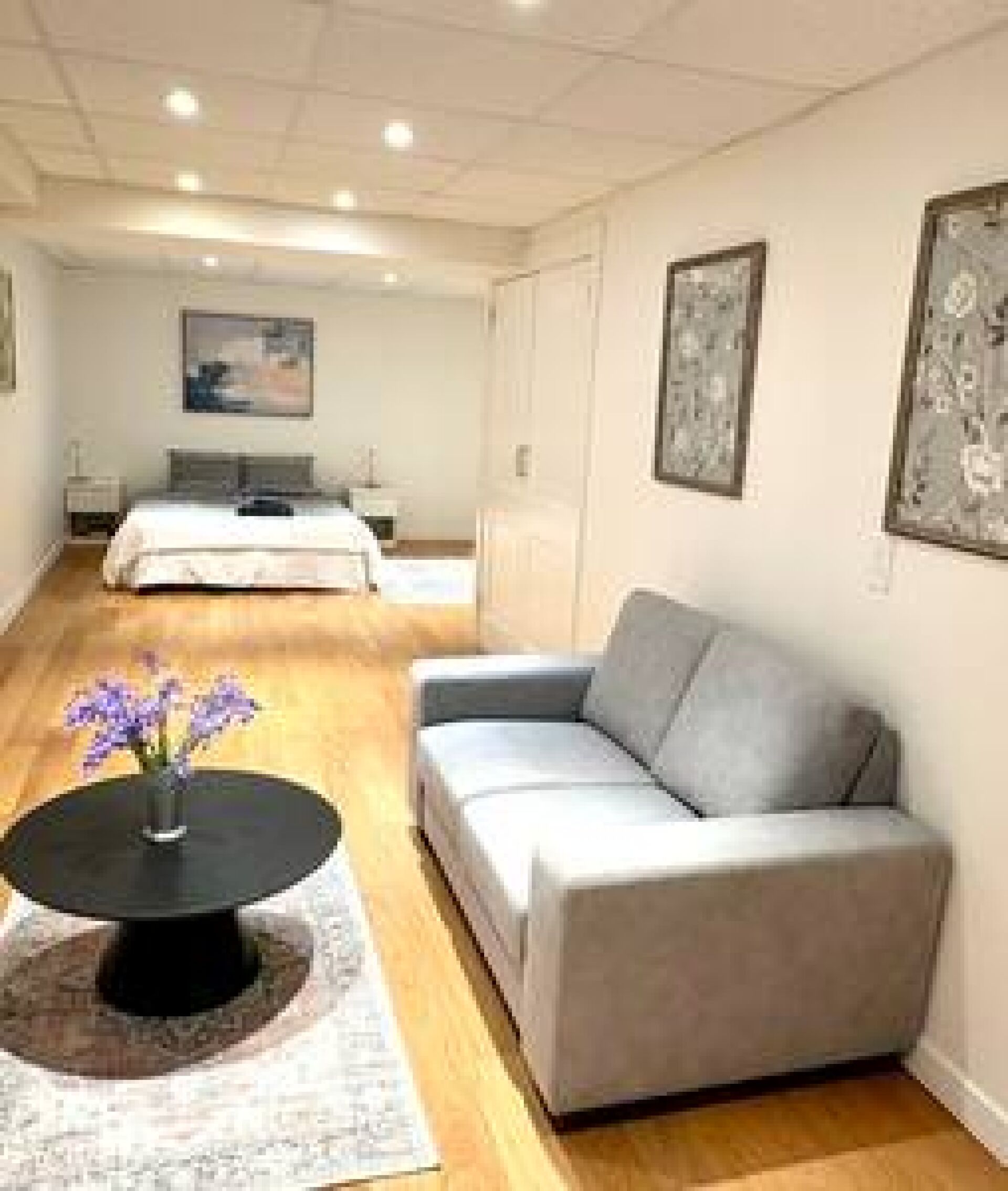 Una habitación de hotel moderna con un sofá gris, una mesa redonda con flores, una cama con edredón blanco y obras de arte enmarcadas en las paredes.