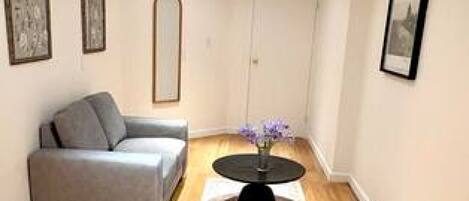 Ein Zimmer mit grauem Sofa, einem runden Tisch mit einer Vase, Holzböden und gerahmten Bildern an den Wänden.